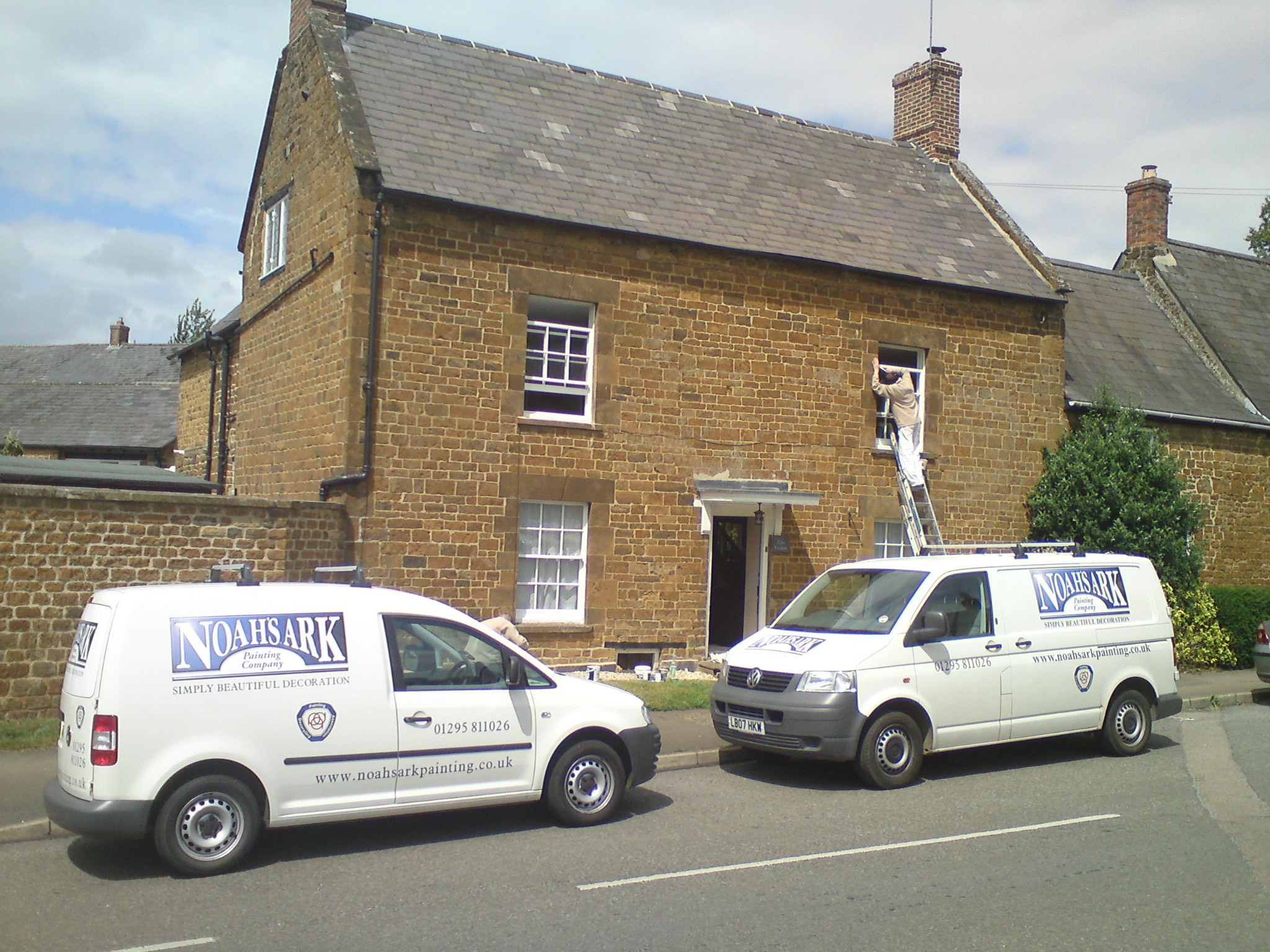 Two Noah's Ark Painting Company vans outside an ironstone cottage while a painter works at a first-floor window